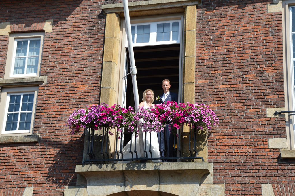 Het bruidspaar op het balkon boven het bordes, achter bloembakken met roze petunia's
